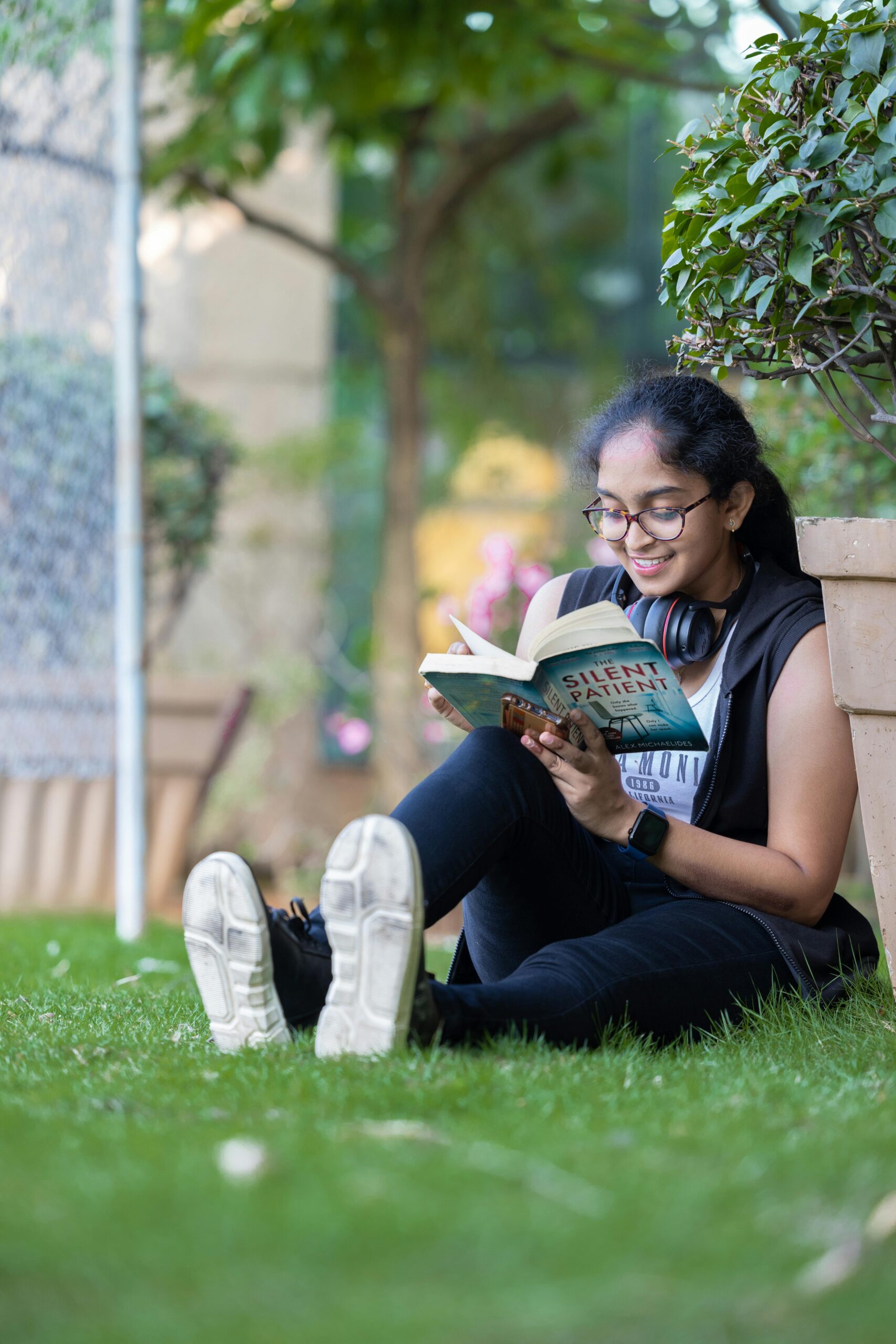 A young woman relaxing in a park, reading a book with headphones around her neck, enjoying a sunny day.